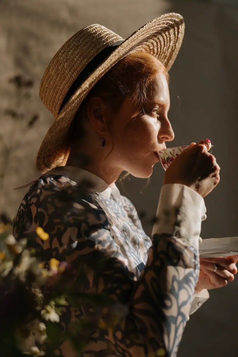 woman in a hat is drinking tea while the sun is beaming in on her