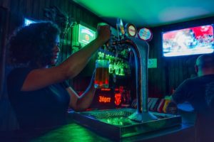 woman filling up a glass of beer in a bar with a man watching tv in the background