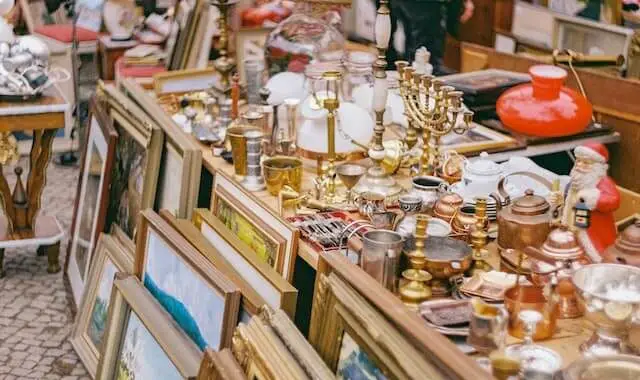 table of miscellaneous goods for sale on a table with paintings in frames on the floor at a flea market