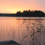 lake with a dock in the foreground and trees and sunset in the background