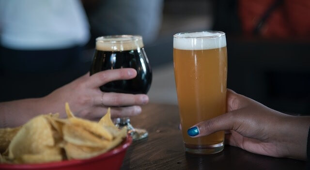 2 hands holding 2 different types of beer with chips in the foreground